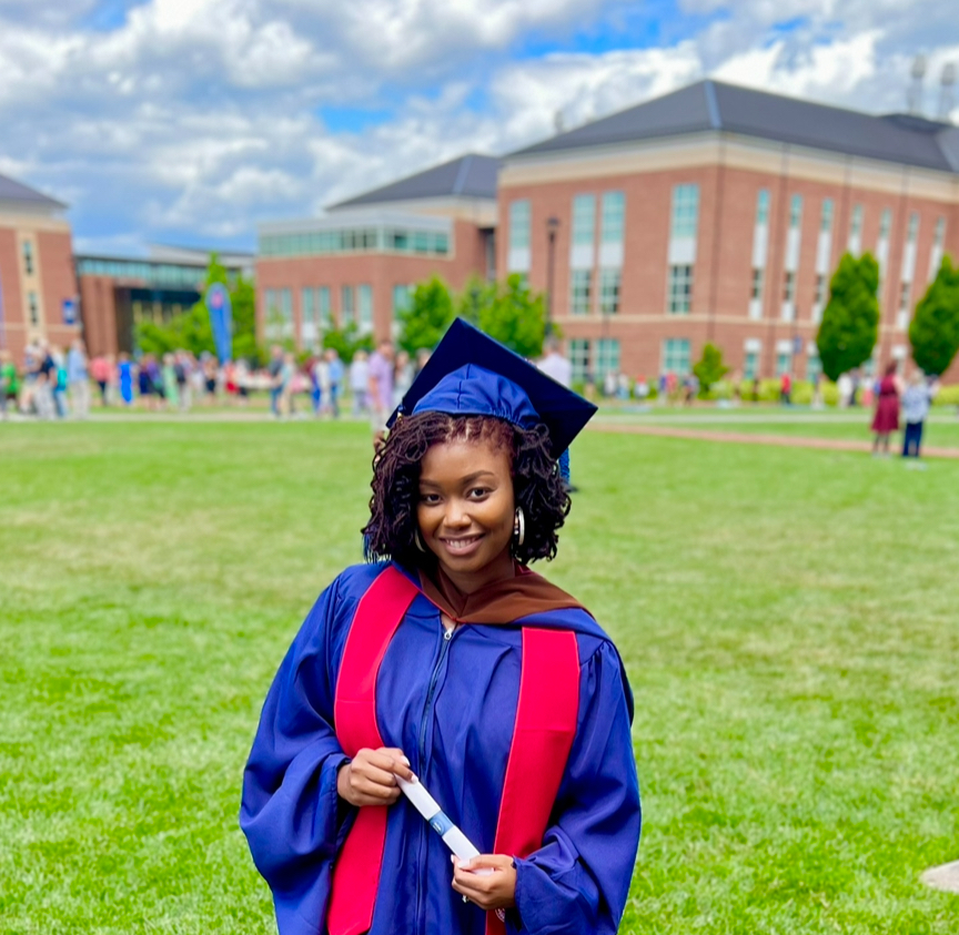 Brenisha in full graduation attire on campus grounds with university buildings in the background, marking the culmination of her educational journey