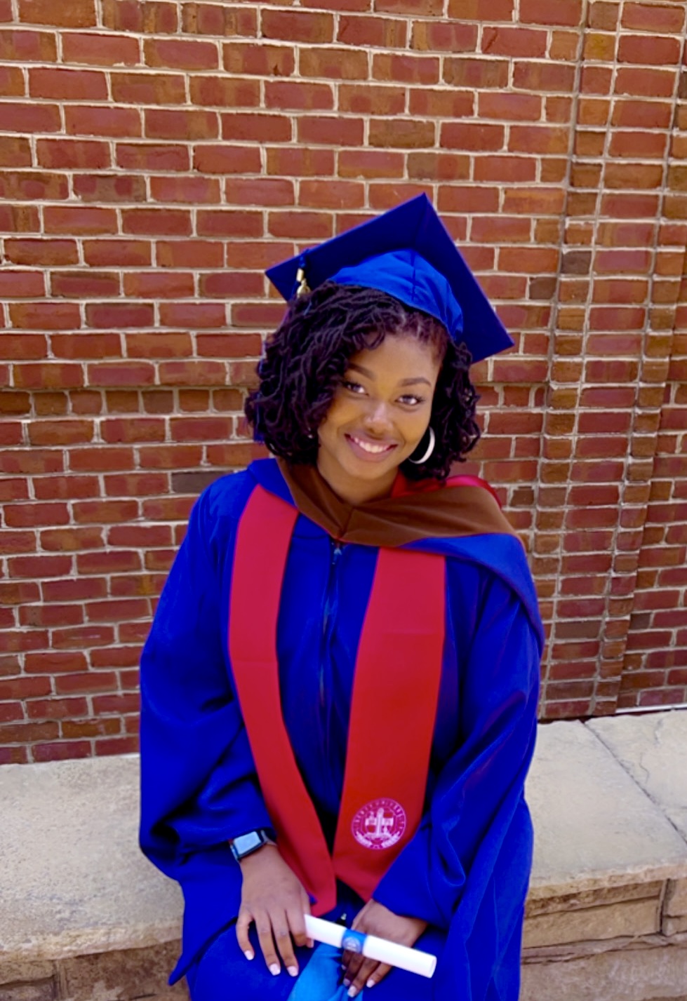 Brenisha in blue graduation regalia with red stole, holding her diploma against a brick wall backdrop, representing her academic achievement in Human-Centered Design